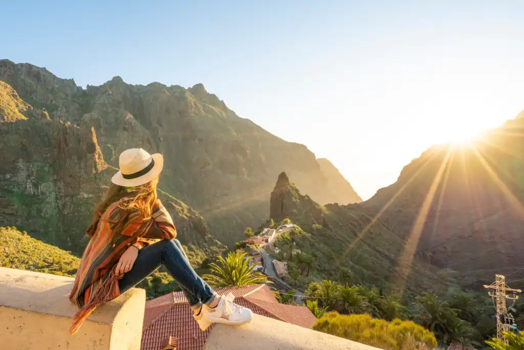 A person wearing a hat and poncho sits on a stone ledge, looking at a sunlit mountainous landscape with a small road and lush greenery in the valley below. Sun rays shine through the peaks.