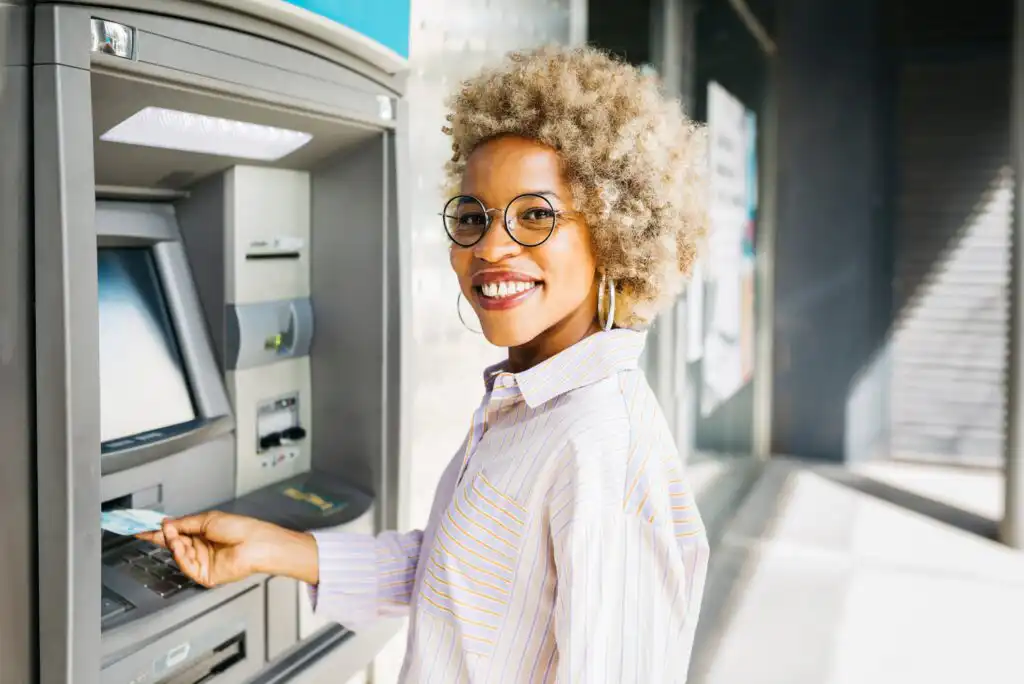 A smiling woman with curly blonde hair and glasses is inserting a bank card into an outdoor ATM. She is wearing a striped shirt and looking back at the camera.