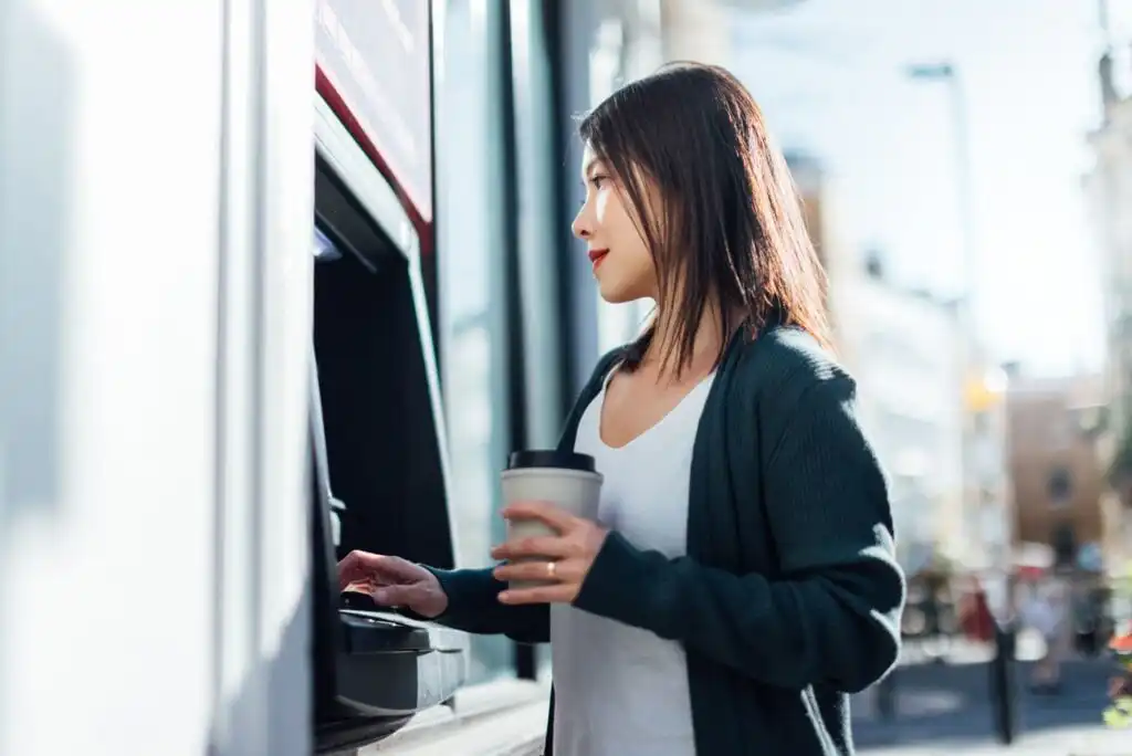 A woman holding a coffee cup uses an outdoor ATM during the day. She is wearing a dark cardigan and white shirt, and appears focused on the ATM screen. The background is a blurred urban street scene.