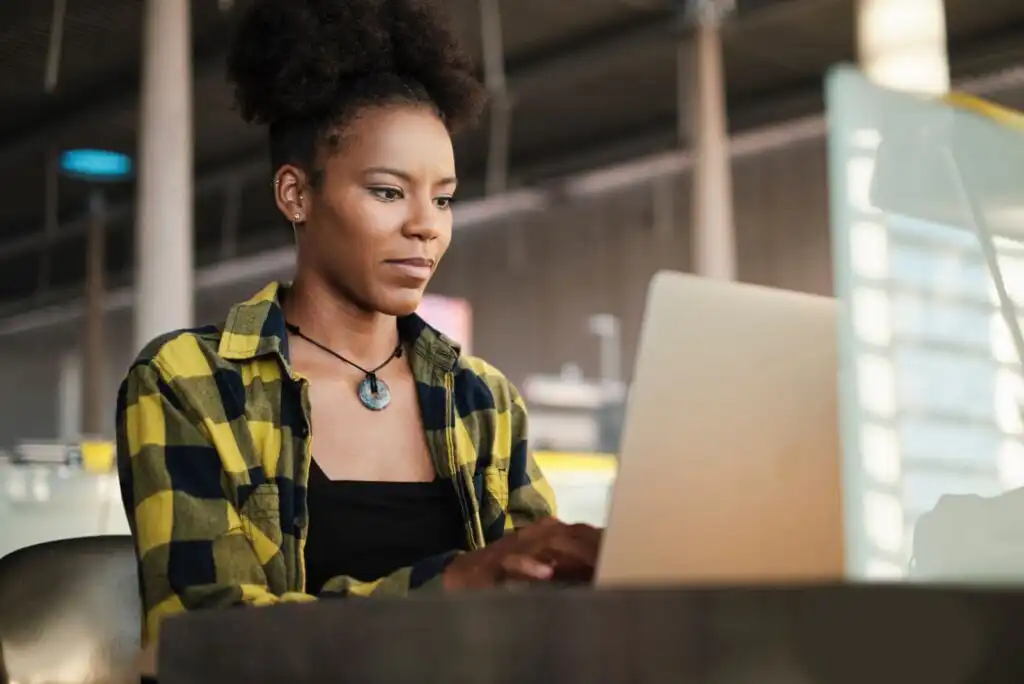 A woman wearing a yellow and black plaid shirt and necklace works on a laptop at a table in a modern, well-lit indoor space. She appears focused on her task.