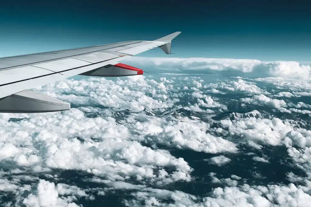 View from an airplane window showing the plane’s wing and a vast expanse of fluffy white clouds below, with mountain peaks barely visible through the clouds and a blue sky above.