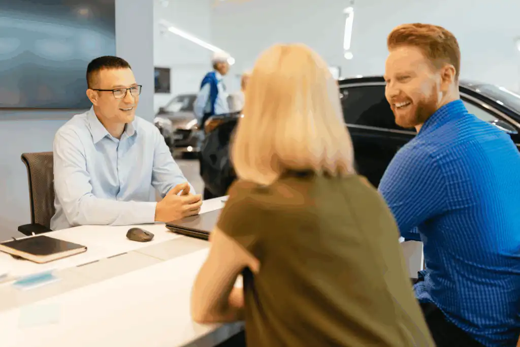 A smiling car dealership employee talks with a man and woman seated across the desk, suggesting a friendly conversation about purchasing a vehicle in a modern showroom.