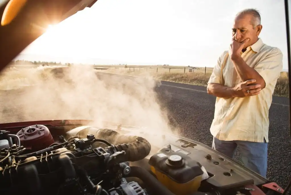 An older man stands by his car on a rural road, looking worriedly at steam rising from the engine under the open hood, with fields and sunlight in the background.