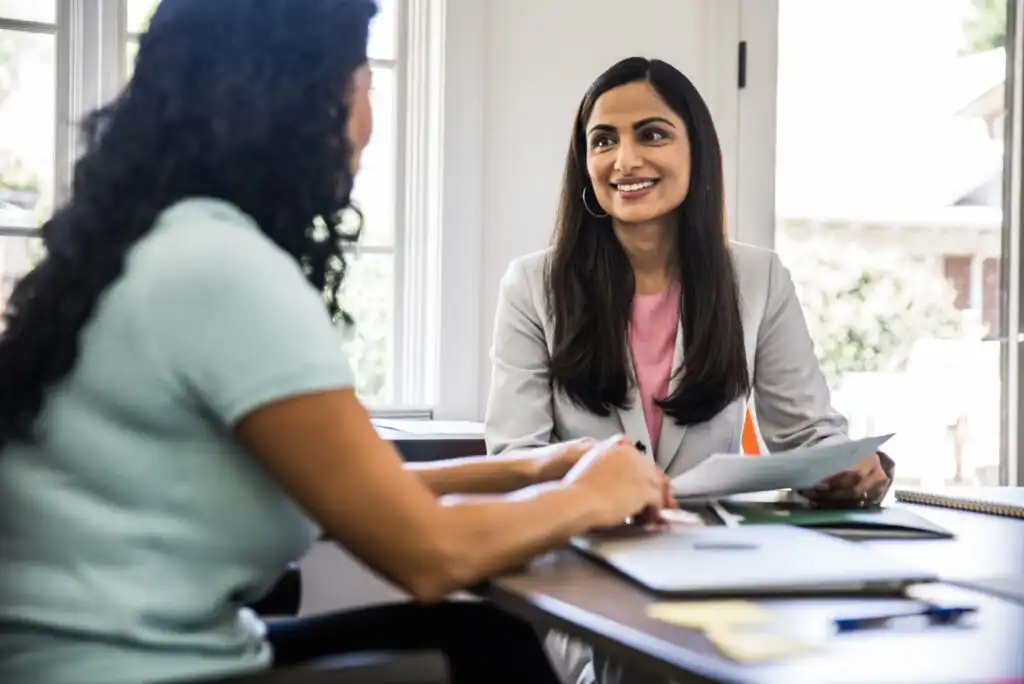 Two women sit at a table in a bright office, engaged in a conversation. One woman is smiling and holding papers, while the other is listening with a laptop and documents in front of her.