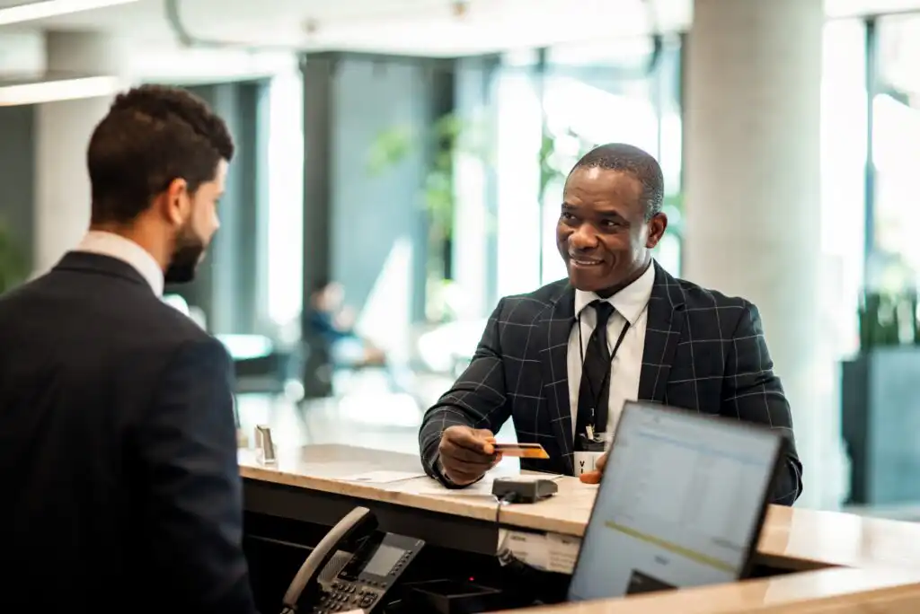 A smiling hotel receptionist in a suit hands a key card to a guest at the front desk, with a computer and phone visible on the counter.