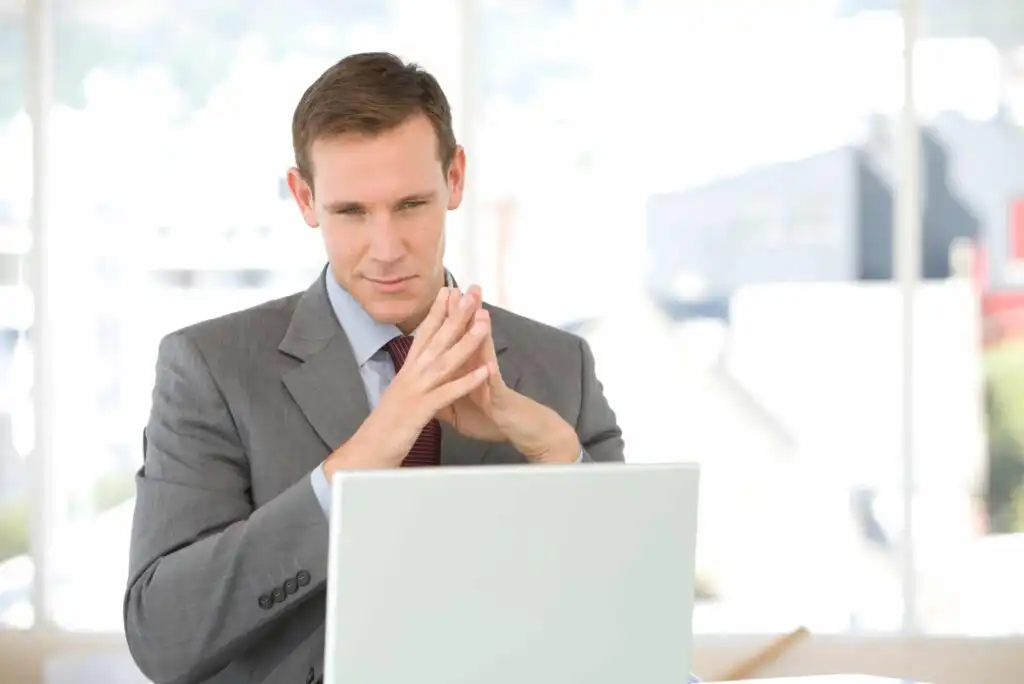A man in a suit sits at a desk, looking thoughtfully at a laptop screen with his hands clasped together. The background is bright and out of focus, suggesting an office environment.