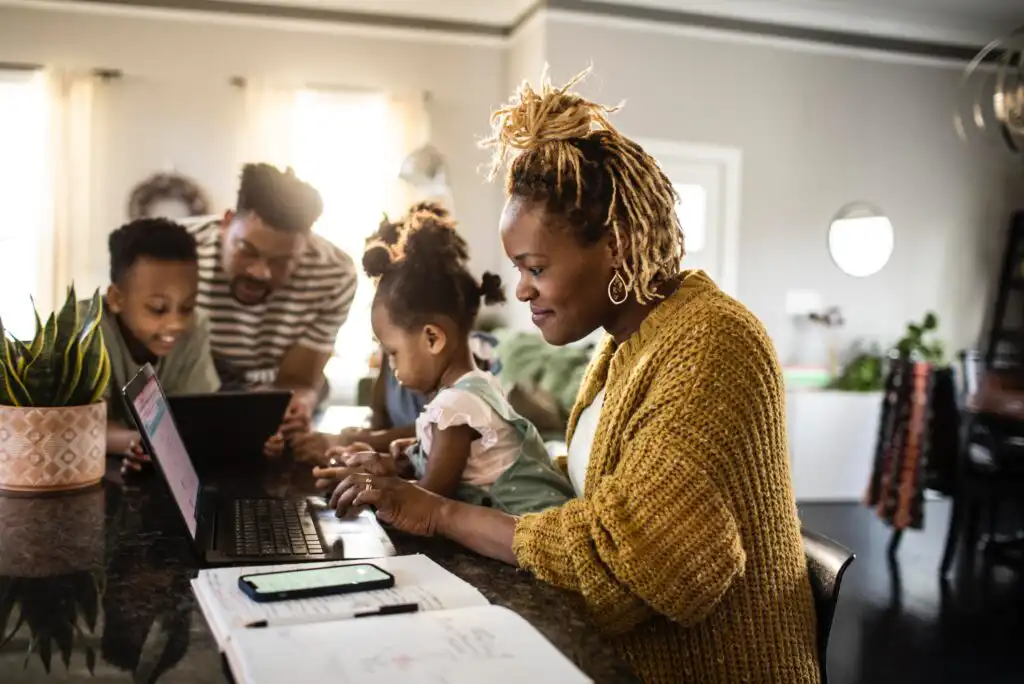 A woman works on a laptop at a kitchen counter with a young girl on her lap, while a man and boy interact in the background. Papers and a phone are on the counter. The family appears engaged and focused.