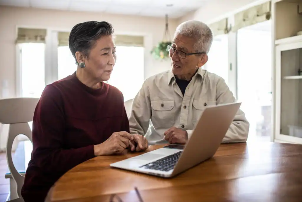 An elderly couple sits at a wooden table, looking at a laptop screen together in a bright, cozy room. The woman wears a red sweater, and the man wears glasses and a beige shirt.