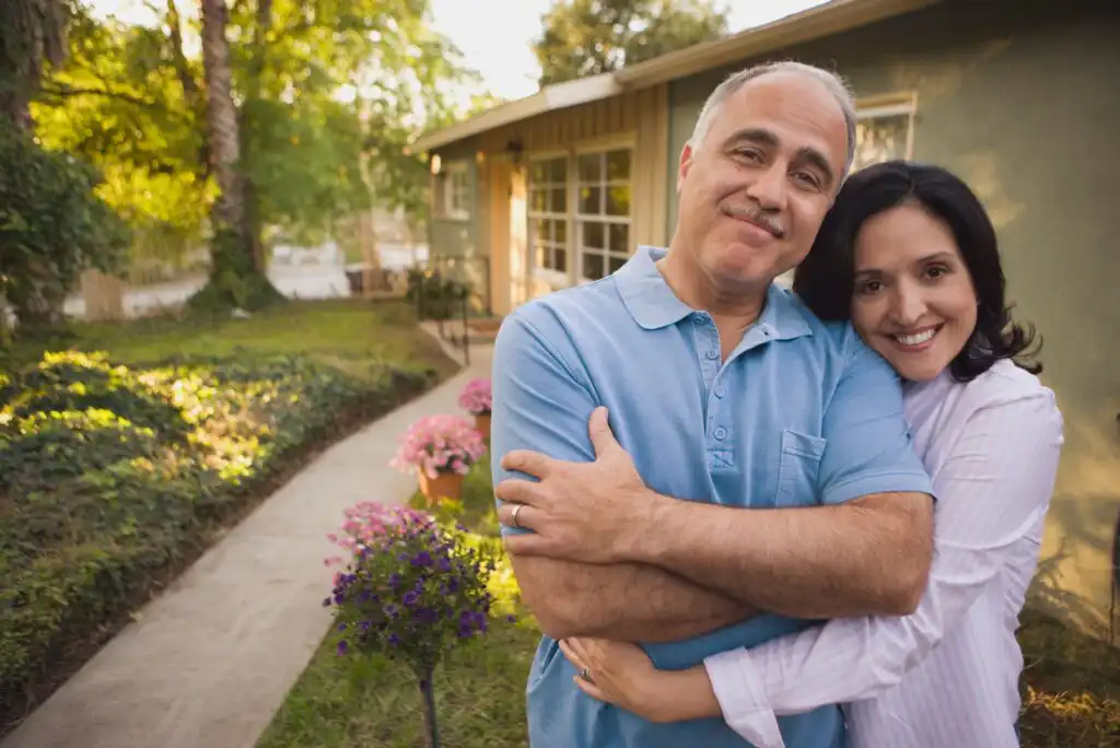 A middle-aged couple smiles and embraces in front of a house with a garden path and blooming flowers, surrounded by lush greenery on a sunny day.