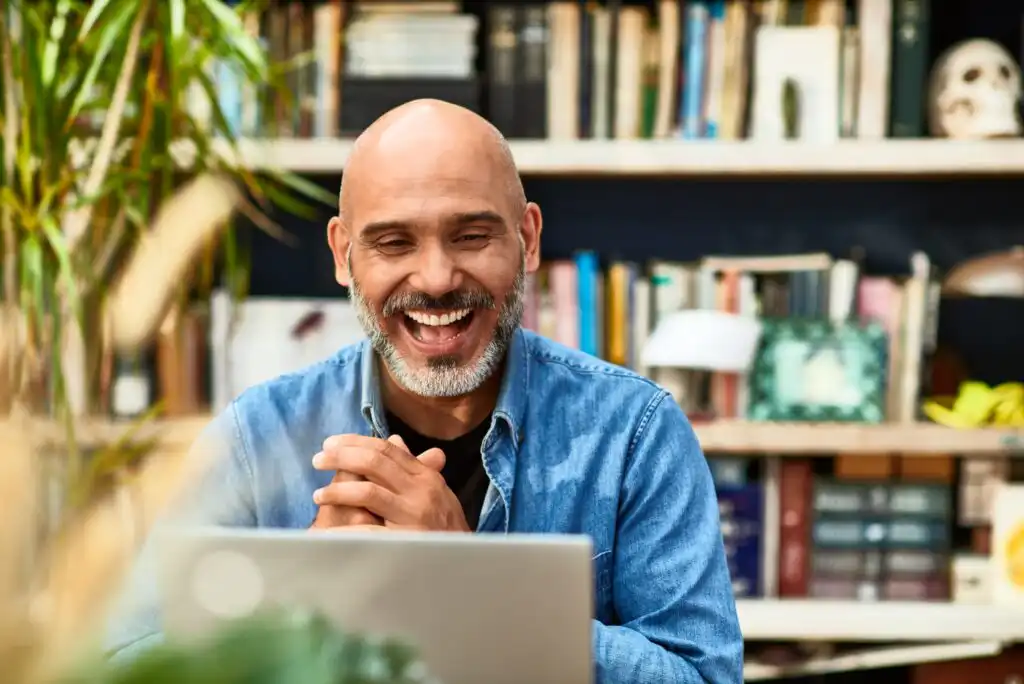 A smiling man with a beard sits at a desk, looking at a laptop. Bookshelves filled with books and decor are in the background, creating a cozy, home office atmosphere.