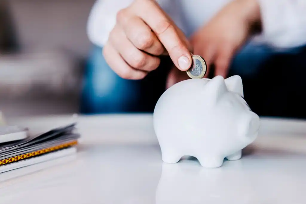 A person’s hand places a coin into a white piggy bank on a table, with a stack of papers or notebooks nearby in the background.