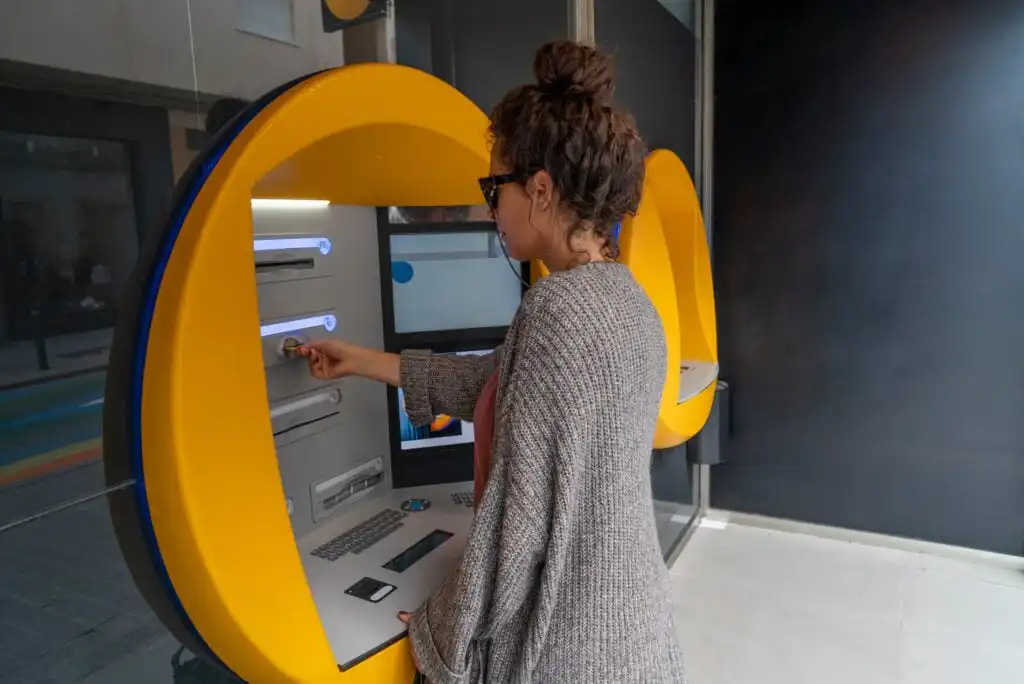 A woman wearing sunglasses and a gray sweater uses an ATM with a bright yellow frame indoors. She is inserting or withdrawing a card from the machine.