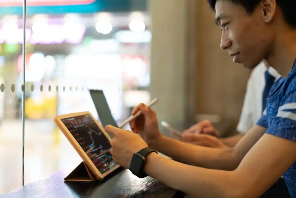 A person using a digital tablet with a stylus, analyzing colorful financial charts on the screen, while sitting indoors next to another individual with a laptop.