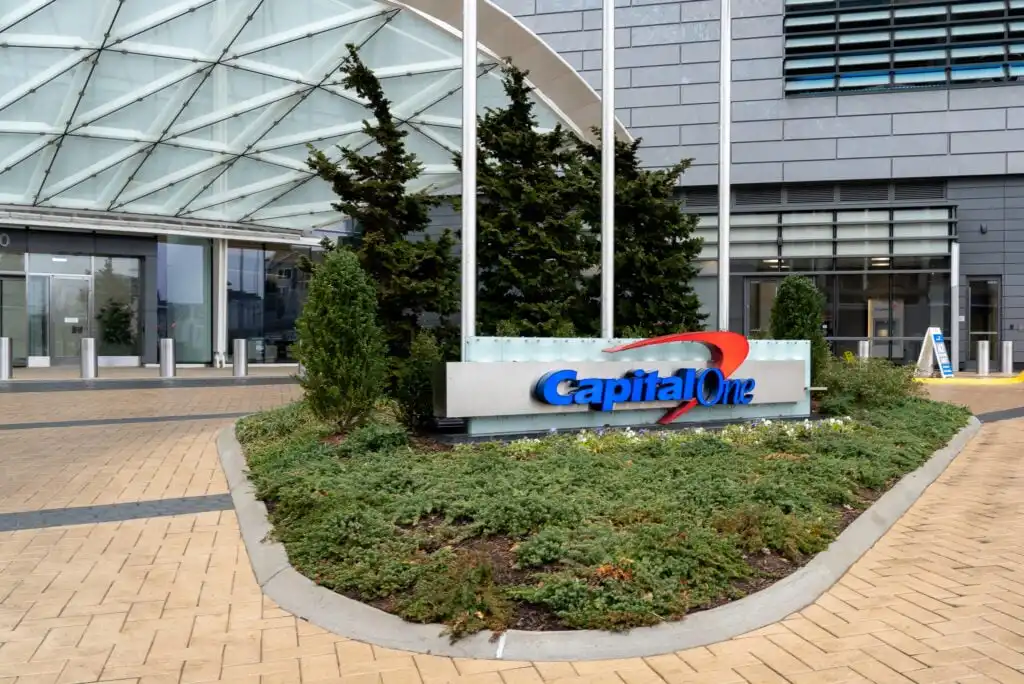 The exterior of a modern building with a glass canopy and a sign that reads Capital One surrounded by greenery and flagpoles.