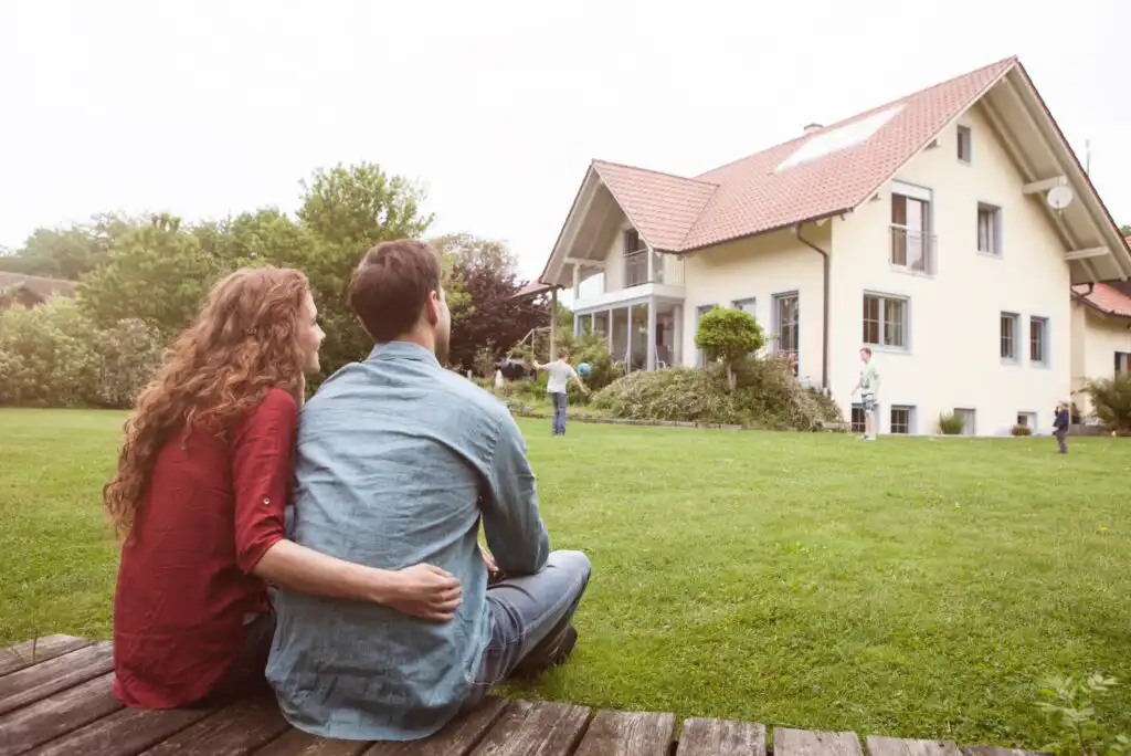 A couple sits on a wooden deck, arms around each other, looking at a modern house with a red roof. Two children play on the lawn, while an adult stands near the house in a green, spacious yard.