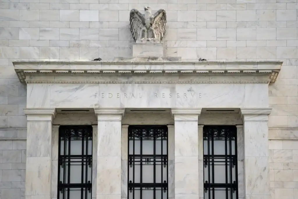 The image shows the exterior of a Federal Reserve building with three tall windows and an eagle sculpture on top of the marble facade, above the engraved words Federal Reserve.