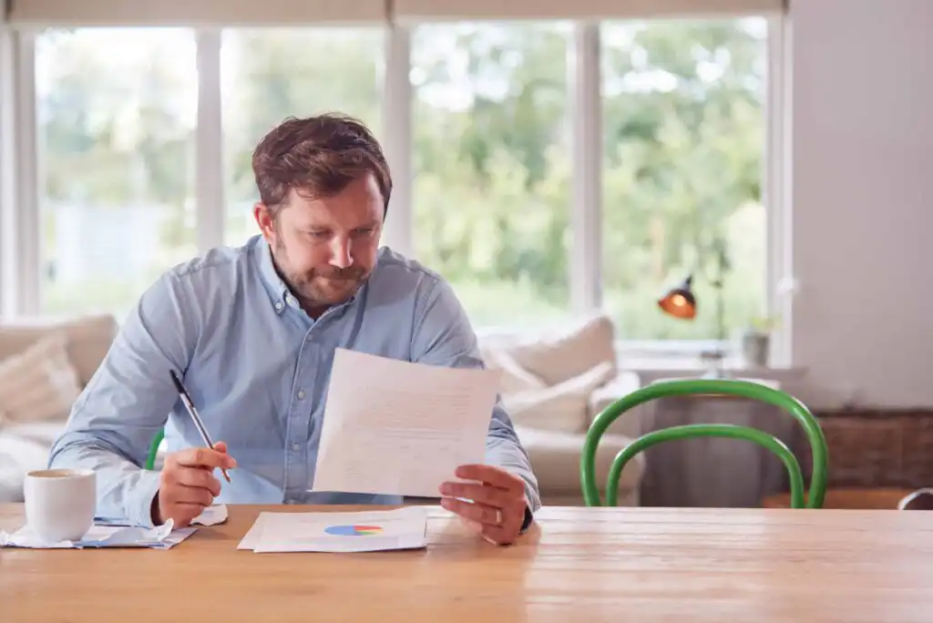 A man sits at a wooden table, reading a document and holding a pen. A coffee mug and papers with charts are in front of him. Large windows and green chairs are visible in the background.