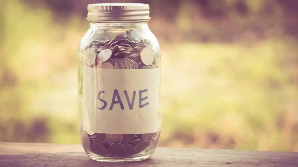 A glass jar filled with coins sits on a wooden surface. A label on the jar reads SAVE in bold letters. The background is softly blurred with natural, outdoor light.