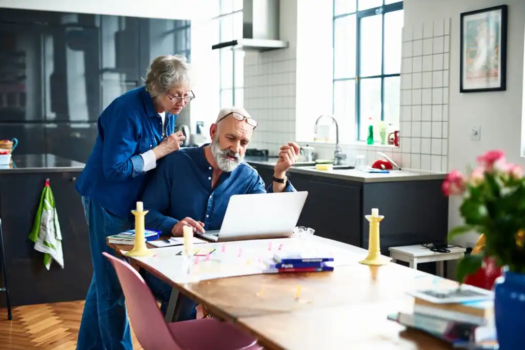 An older couple in a modern kitchen looks at a laptop on a table. The man sits and uses the computer, while the woman leans over his shoulder. The table is cluttered with candles, papers, and books.