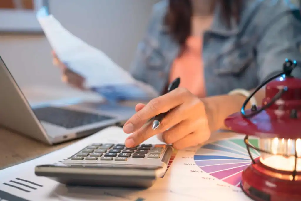 A person using a calculator with one hand while holding papers in the other, sitting at a desk with a laptop, charts, and a red lantern. The focus is on the persons hands and the calculator.