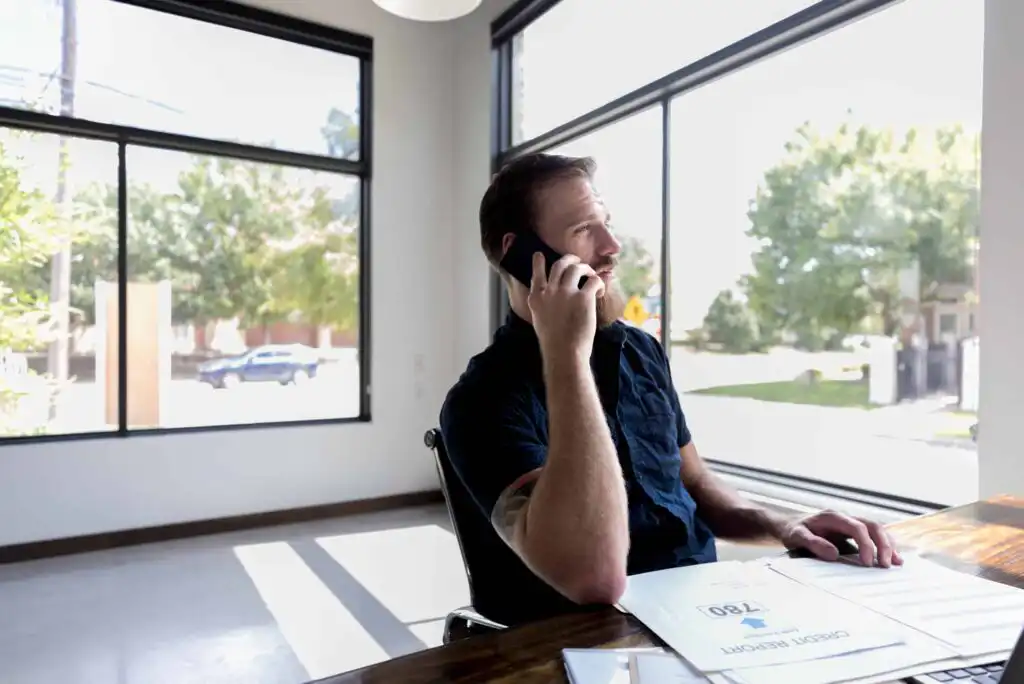 A man sits at a desk by large windows, talking on his phone and looking outside. Sunlight fills the modern office, and papers, including a credit report, are on the desk in front of him.