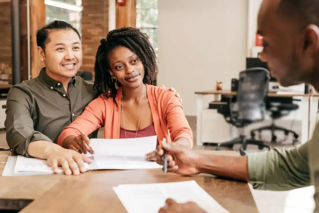 A smiling couple sits at a desk holding documents, attentively listening to a person across from them, who is gesturing with a pen. The setting appears to be a bright, modern office.