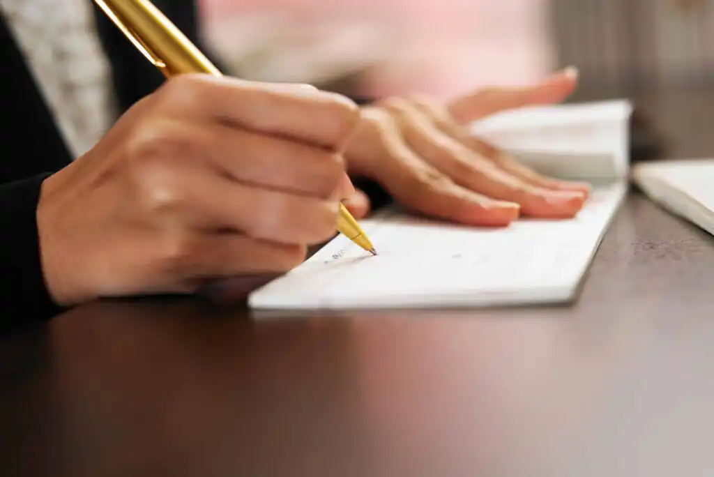 A person writes on a check with a gold pen, resting one hand on the checkbook and the other hand on the table. The focus is on the hands and pen, with a blurred background.
