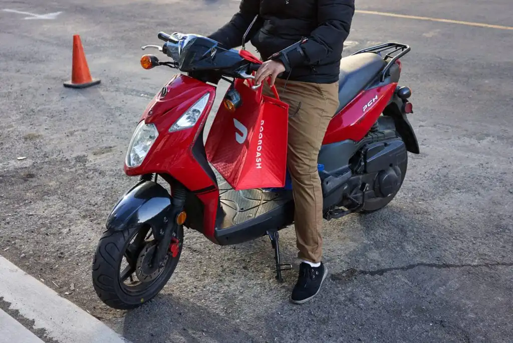 A person in a black jacket and tan pants sits on a red scooter, holding a red DoorDash delivery bag while parked on a street near an orange traffic cone.