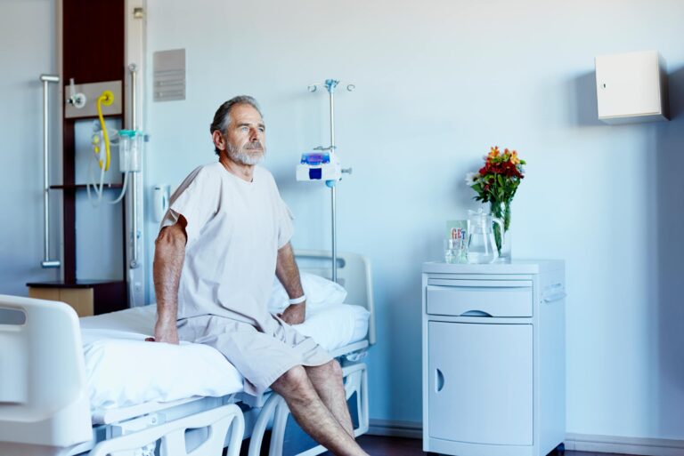 Thoughtful Mature Man Looking Away While Sitting On Bed In Hospital Ward