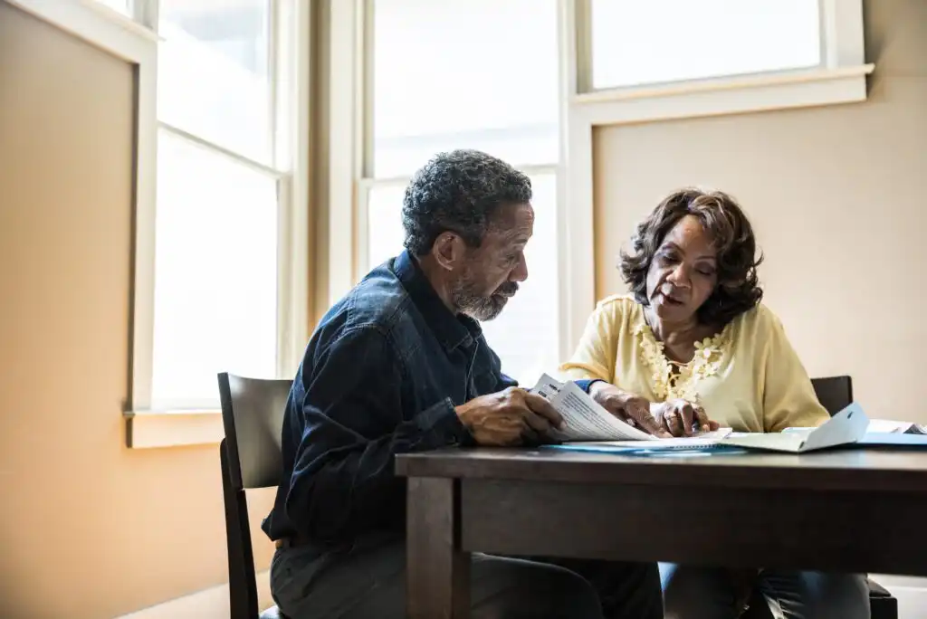 An older couple sits at a table by large windows, reviewing and discussing documents together. The woman points at a paper while the man listens attentively. Both appear focused and engaged.