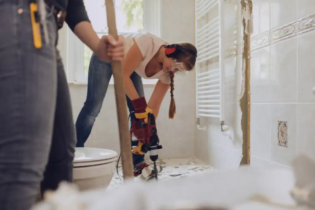 A person wearing goggles and earmuffs uses a power tool to break up a tile floor in a bathroom, while another person holds a tool nearby. Debris is scattered on the floor.