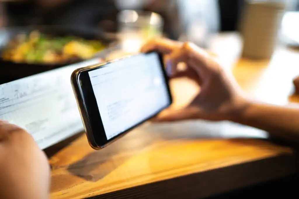 A person holds a smartphone and takes a photo of a check or bill on a wooden table in a restaurant, with blurred food and cups in the background.