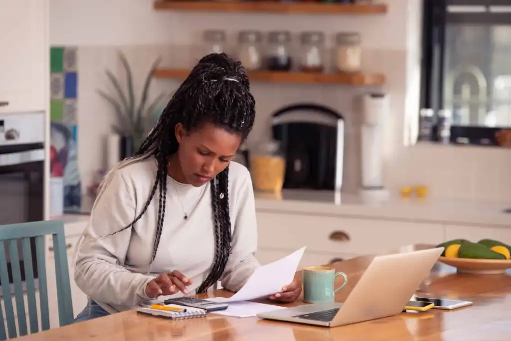 A woman sits at a kitchen table, looking at documents and using a calculator. A laptop, notebook, mug, and phone are on the table. The kitchen is bright, with shelves and various items in the background.