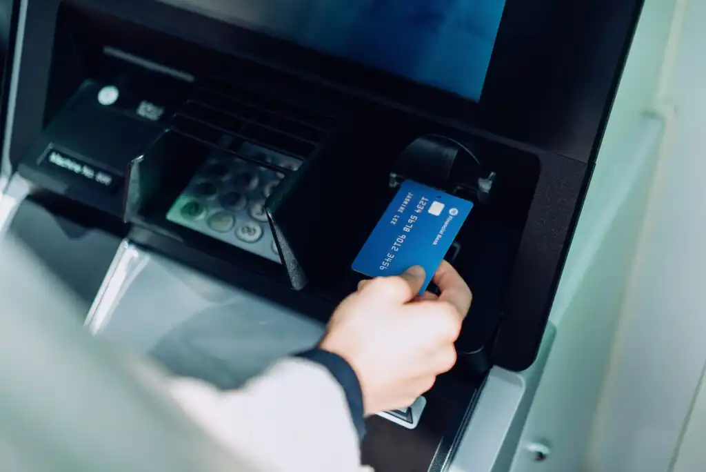 A person’s hand is inserting a blue credit card into an ATM machine, with a keypad and screen visible in the background.
