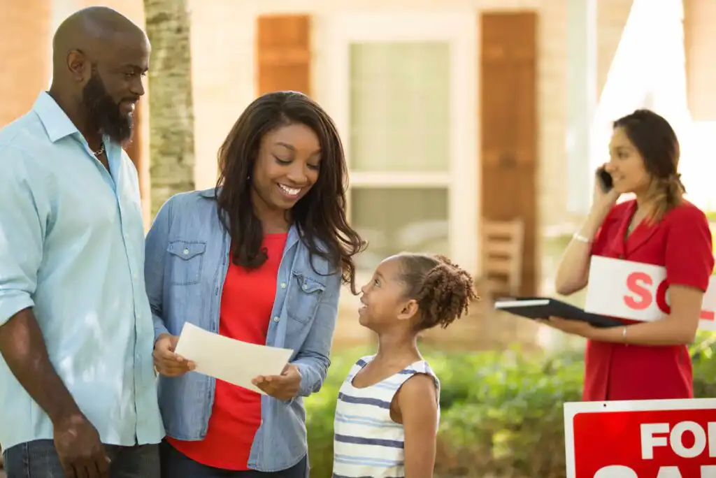 A smiling family stands outside a house with papers in hand while a young girl looks up at her mother. In the background, a real estate agent talks on the phone next to a For Sale sign.