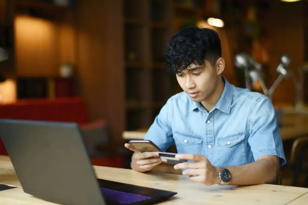 A young man in a denim shirt sits at a wooden table with a laptop, holding a smartphone and a credit card, appearing to make an online purchase in a cozy, softly lit room.