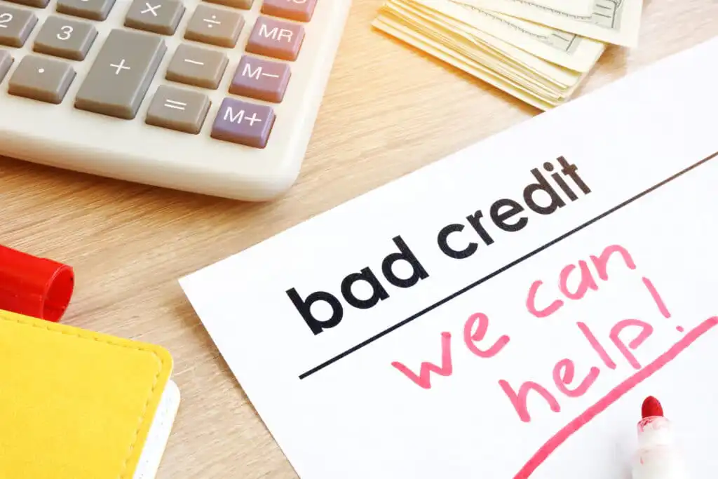 A paper with bad credit printed and we can help! written in pink marker, next to a calculator, a stack of cash, and a red marker on a wooden desk.