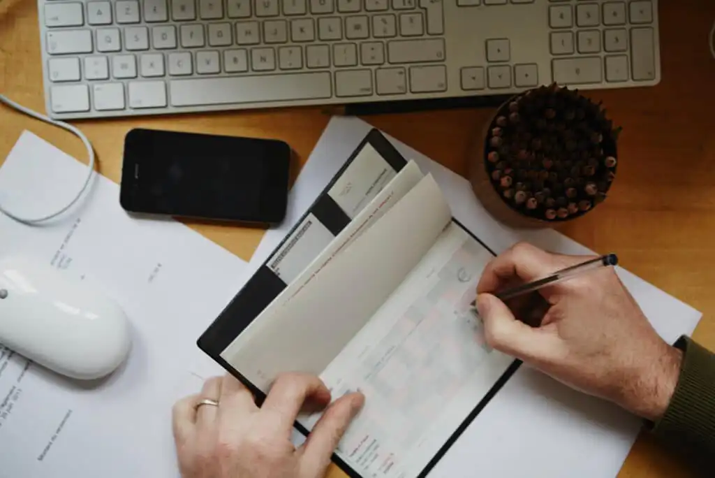 A person writes in a checkbook on a desk with a keyboard, mouse, phone, papers, and a bowl of colored pencils.