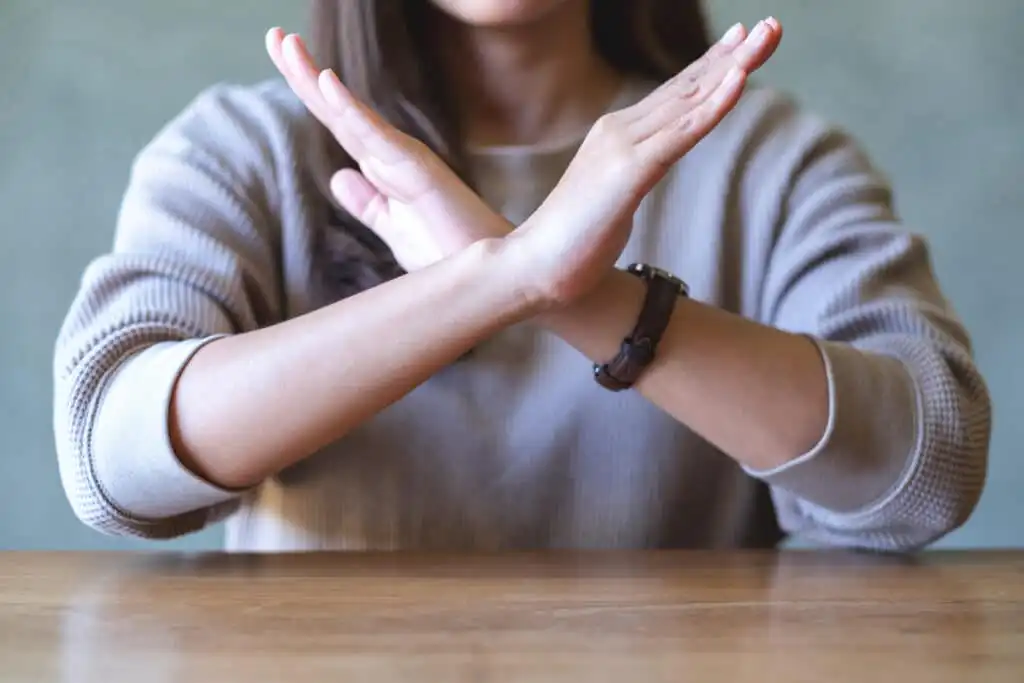 A person sitting at a table with arms crossed in front of their chest, forming an X gesture, suggesting refusal or prohibition. The persons face is partially out of frame.