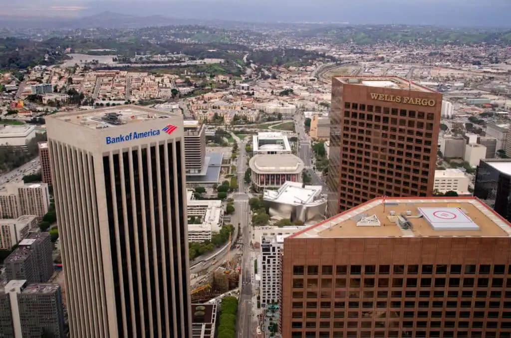 Aerial view of downtown Los Angeles showing tall Bank of America and Wells Fargo buildings in the foreground, with city streets, smaller buildings, and green hills in the background.