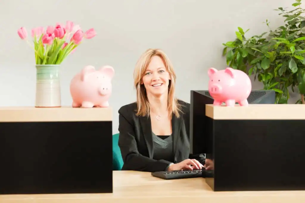 A smiling woman in business attire sits at a desk with a computer keyboard, flanked by two pink piggy banks. A vase with pink tulips and a green plant decorate the workspace.
