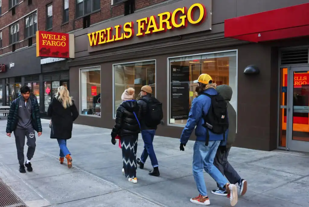 People walk past a Wells Fargo bank branch on a city sidewalk, with the bank’s large yellow and red sign visible above the entrance and several windows displaying the interior.