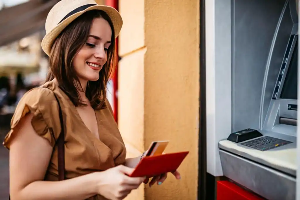 A young woman wearing a tan dress and straw hat smiles while holding a wallet and bank card, standing next to an outdoor ATM machine.
