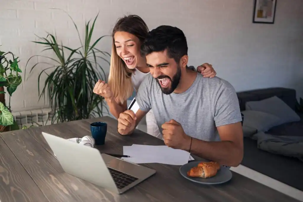 A smiling man and woman sit at a table with a laptop, celebrating and cheering with raised fists. Papers, a coffee mug, and a plate with a croissant are on the table. They appear happy and excited by something on the screen.