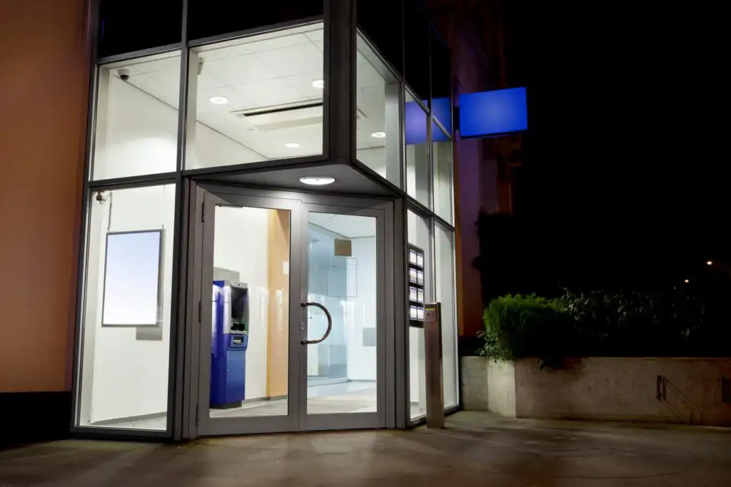 A glass-fronted bank entrance at night, brightly lit inside, with an ATM visible on the left and a blue blank sign above the door. Shrubs and a concrete planter are outside.