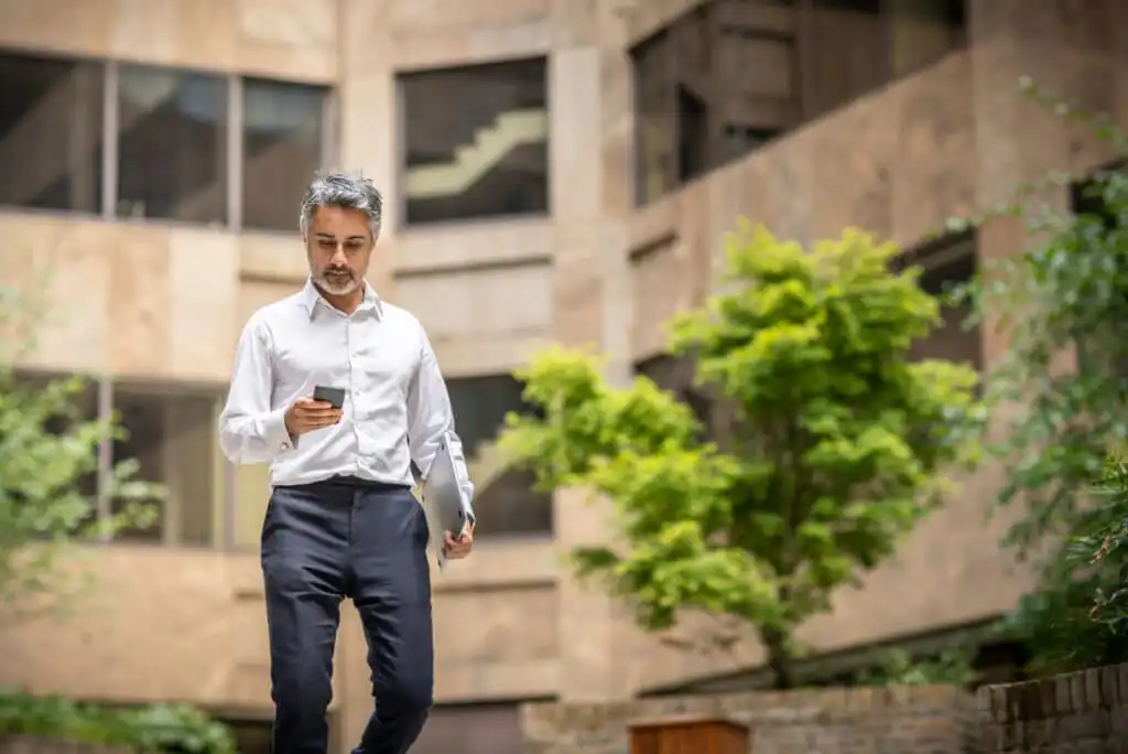 A man in business attire walks outdoors in an urban area, looking at his smartphone while holding a laptop. Green trees and a stone building are in the background.