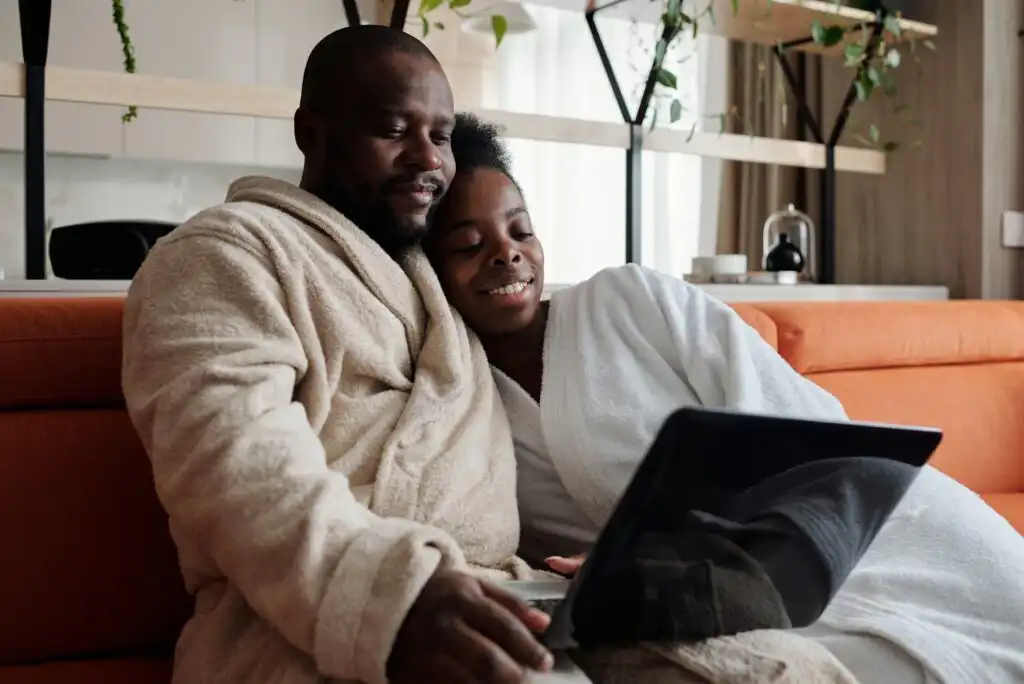A couple wearing bathrobes sits close together on a couch, smiling and looking at a laptop screen. They appear relaxed and comfortable in a cozy home setting with plants in the background.