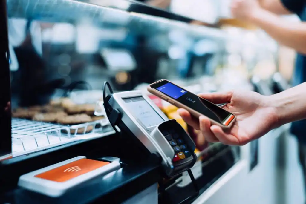 A person holds a smartphone over a contactless payment terminal at a counter, making a digital payment. The background is blurred, showing food on display and other people.
