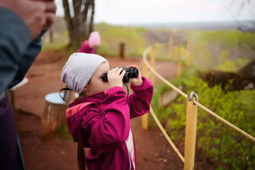 A young child in a pink jacket and white hat looks through binoculars outdoors on a dirt path, surrounded by greenery and a yellow rope fence, with another person blurred in the background.