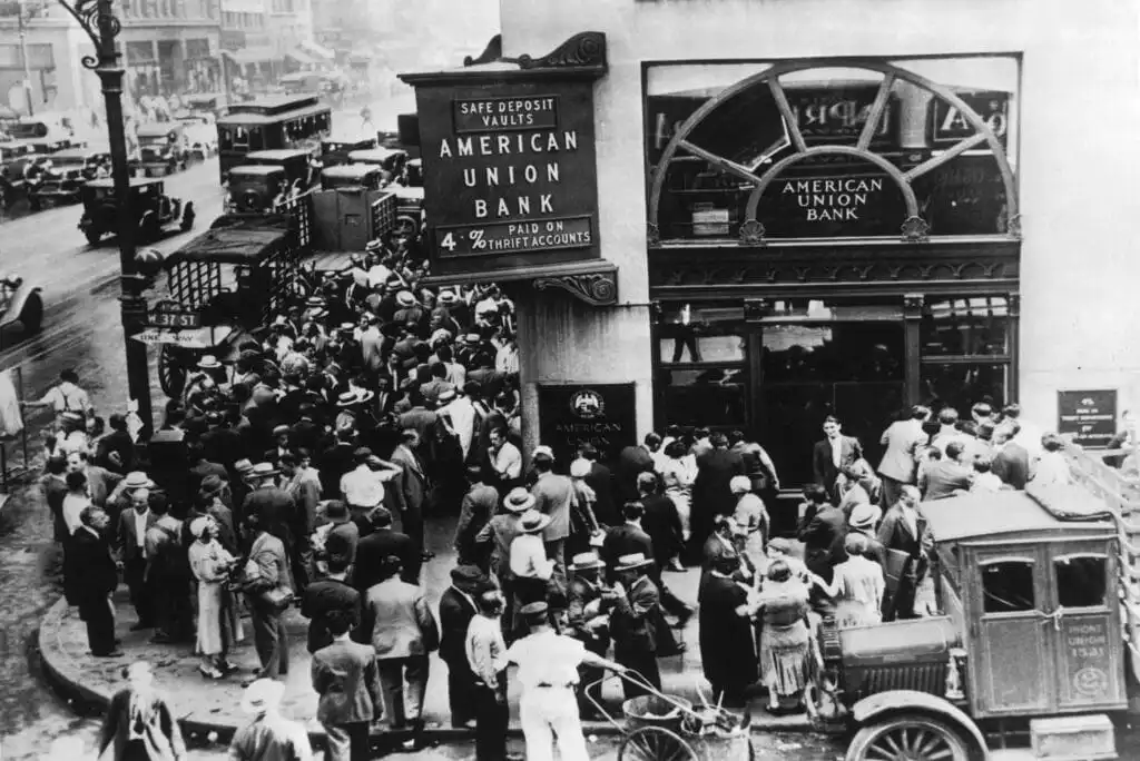 A large crowd gathers outside the American Union Bank during the Great Depression, with people lining up on the street near parked cars, seeking to withdraw their money amid financial panic.